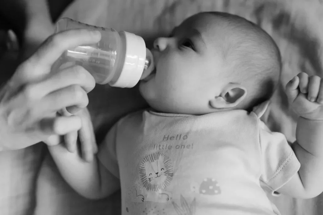 A baby being fed with a bottle in a black and white photograph while lying down.